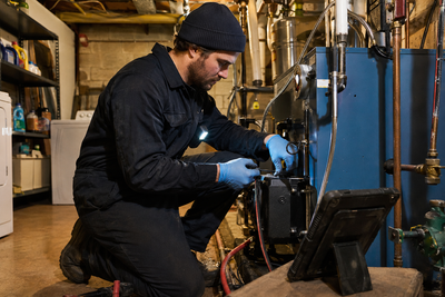 A heating service technician servicing an oil-fired boiler in a basement, a rugged tablet nearby.